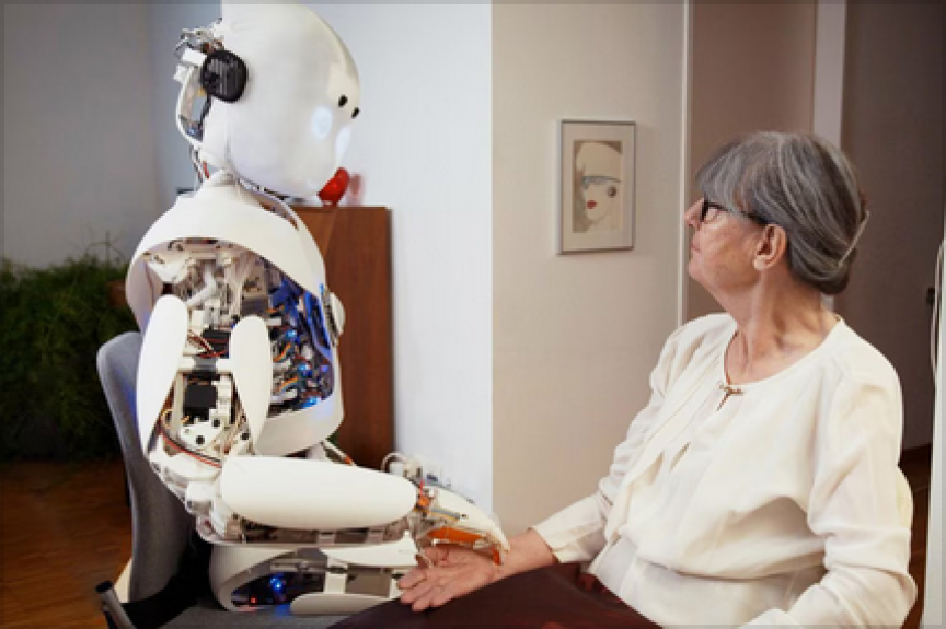 Ältere Frau mit Brille sitzt einem humanoiden Roboter gegenüber und berührt dessen Hand an einem Tisch.