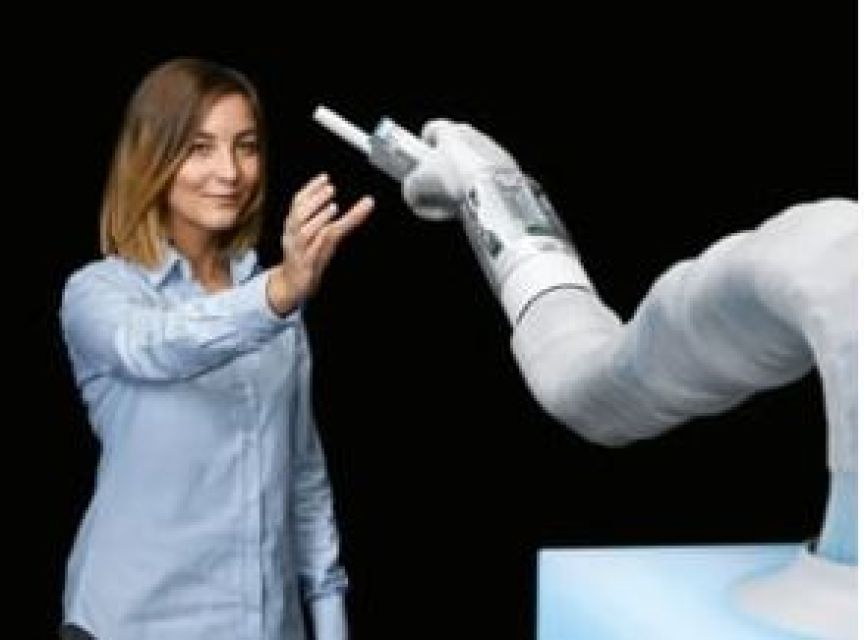 Frau in blauem Hemd interagiert mit einem weißen Roboterarm, der einen Stift hält. Schwarzer Hintergrund.